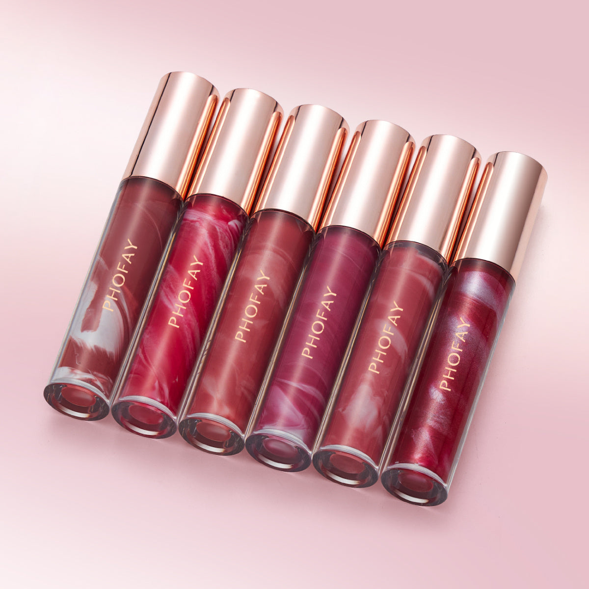 Six tubes of PHOFAY Rose Blooming Hydrating Lip Gloss by PHOFAY Luxe Cosmetics, with rose gold caps, are arranged diagonally on a pink surface in shades of red, pink, and mauve. Moisturizing ingredients keep lips soft and nourished all day.